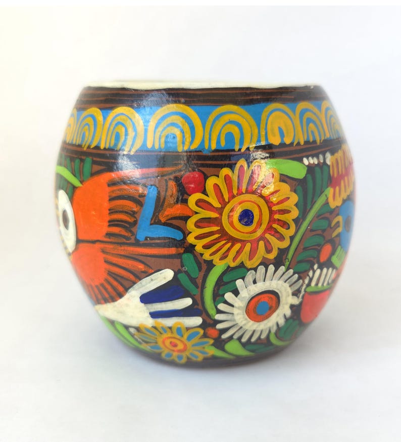 May include: A hand-painted, round, brown ceramic vessel with a white rim. The vessel is decorated with colorful floral and bird designs in orange, yellow, blue, and green. The vessel is likely used for decorative purposes.