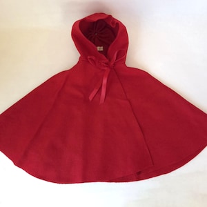 Puede incluir: Una capa con capucha de color rojo brillante confeccionada en un tejido liso y de color sólido. La prenda incluye una capucha integrada y un cierre de lazo de cinta roja a juego en el cuello, diseñado para caer holgadamente sobre los hombros en corte circular.