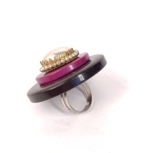 May include: A ring made from layered buttons with a pearl and gold trim. The ring is black, pink, and white.
