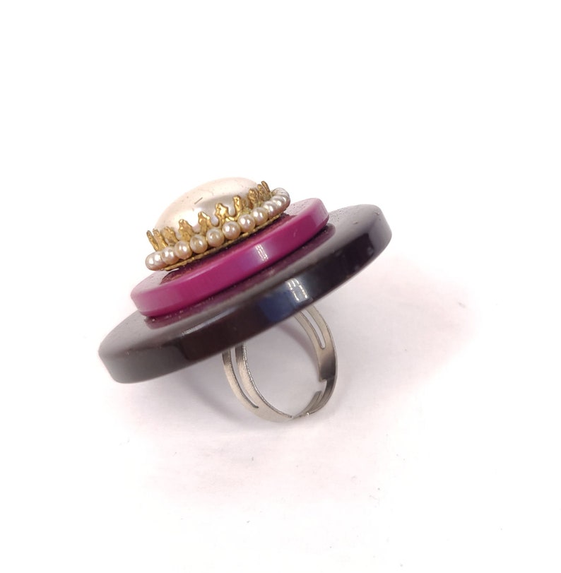 May include: A ring made from layered buttons with a pearl and gold trim. The ring is black, pink, and white.