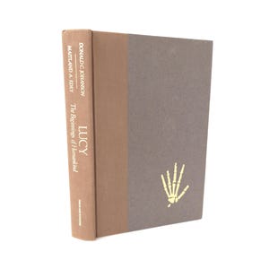 May include: A hardcover book titled "LUCY" by Donald C. Johanson and Maitland A. Edey. The book has a brown spine and a gray cover with a gold skeletal hand illustration. The title "The Beginnings of Humankind" is also printed on the spine.