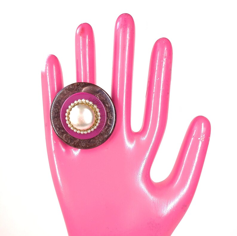 May include: A ring with a brown and pink button-like design. The button has a white pearl-like center and a gold rim.