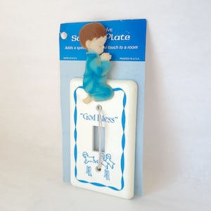 Puede incluir: Una placa de interruptor de luz blanca con un borde azul y un diseño azul y blanco. Una pequeña figura de plástico de un niño rezando está unida a la parte superior de la placa. La placa tiene las palabras "God Bless" impresas en ella.
