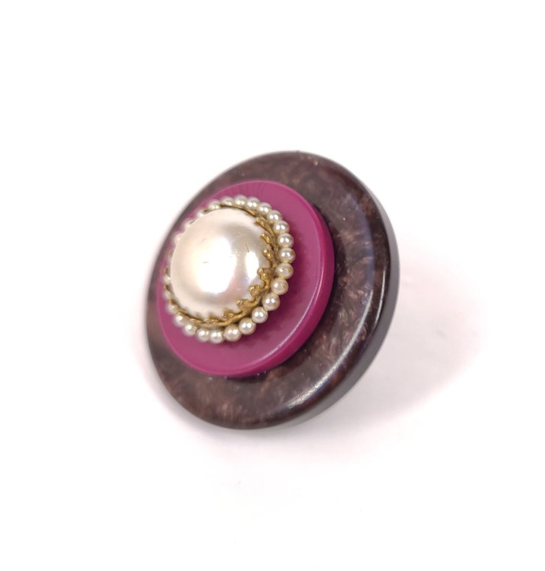 May include: A round, multi-layered button with a faux pearl center. The button has a dark brown outer layer, a pink middle layer, and a white pearl center with a gold trim.