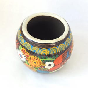 May include: A hand-painted, round ceramic vase with a wide opening. The vase features a brown base with colorful floral and bird designs in orange, yellow, blue, and white. The rim is white.