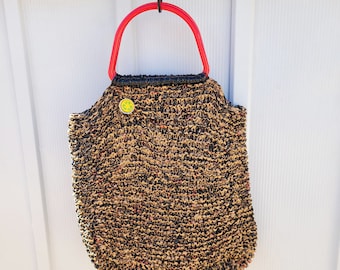 May include: A brown and black woven tote bag with a red plastic handle hanging on a white wall.