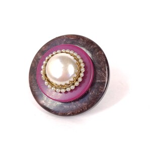 May include: A round, brown and maroon button with a white pearl in the center surrounded by a gold and white beaded trim.