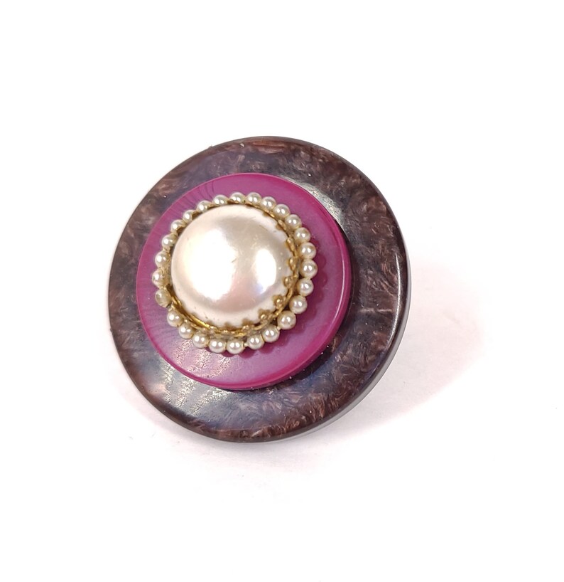 May include: A round, brown and maroon button with a white pearl in the center surrounded by a gold and white beaded trim.