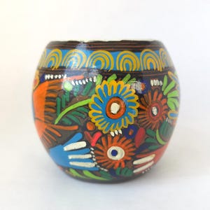 May include: A hand-painted, round, dark brown container with a white rim. The container is decorated with colorful floral and abstract designs in orange, blue, green, and red. The design includes a blue and yellow pattern around the top.