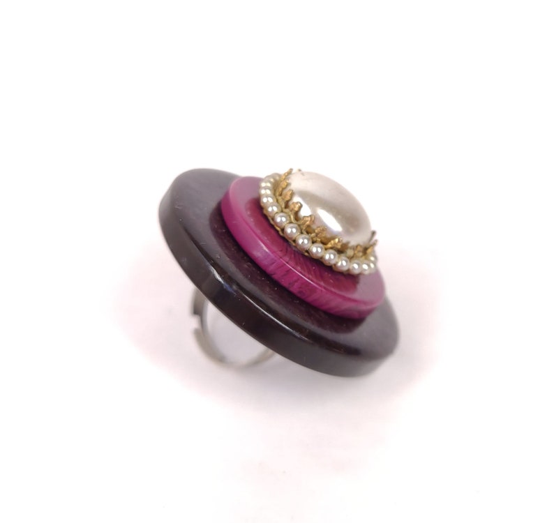 May include: A ring made from layered buttons. The top button is a dark purple with a white pearl and gold trim. The bottom button is black.