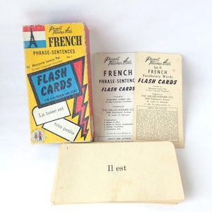 Puede incluir: Juego de tarjetas didácticas francesas vintage. La caja es amarilla, roja y azul con la Torre Eiffel y las palabras "FRENCH FLASH CARDS". El juego incluye una caja, dos folletos y una pila de tarjetas con frases en francés. Las tarjetas son para el aula y el hogar.