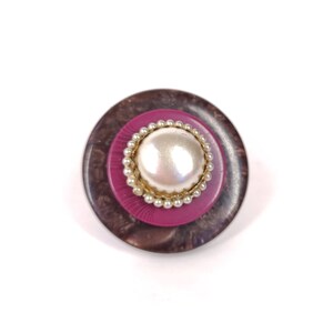 May include: A round, brown and pink button with a white pearl center. The button has a decorative gold rim around the pearl.