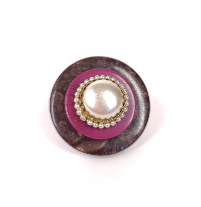 May include: A round, brown and pink button with a white pearl center. The button has a decorative gold rim around the pearl.