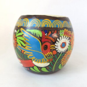 May include: A hand-painted, round, wooden container with a dark brown base and a cream-colored rim. The container features a vibrant design of a blue bird, flowers, and foliage in red, green, yellow, and white. The top border has a blue wave pattern.
