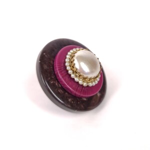 May include: A large, round, two-toned button with a faux pearl center. The button is brown and black on the outside and pink on the inside. The pearl is surrounded by a gold trim and a row of small white beads.