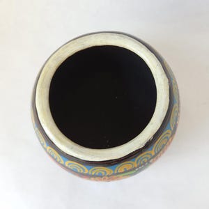 May include: Overhead view of a round, hand-painted ceramic bowl. The interior is black, with a white rim. The exterior features a brown base with a colorful, repeating pattern of blue, yellow, and orange swirls.