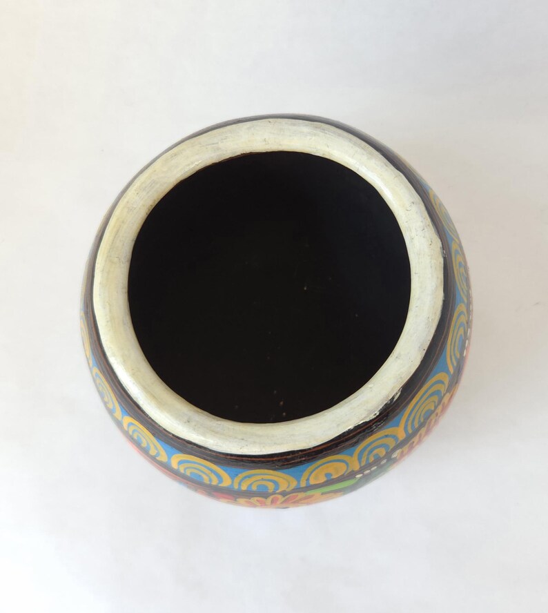May include: Overhead view of a round, hand-painted ceramic bowl. The interior is black, with a white rim. The exterior features a brown base with a colorful, repeating pattern of blue, yellow, and orange swirls.