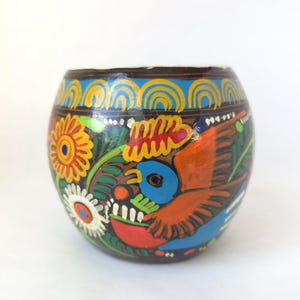 May include: Hand-painted, round decorative container with a dark brown base. The container features a vibrant design of flowers, a bird, and other colorful patterns in orange, yellow, blue, and white. The rim has a blue and yellow design.