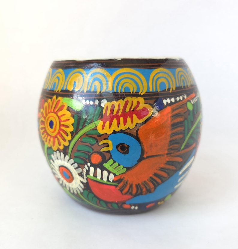 May include: Hand-painted, round decorative container with a dark brown base. The container features a vibrant design of flowers, a bird, and other colorful patterns in orange, yellow, blue, and white. The rim has a blue and yellow design.
