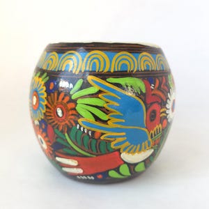 May include: A hand-painted, round wooden container with a dark brown base. The container is adorned with colorful floral and bird designs in blue, green, red, orange, and white. The rim features a blue and yellow arched pattern.