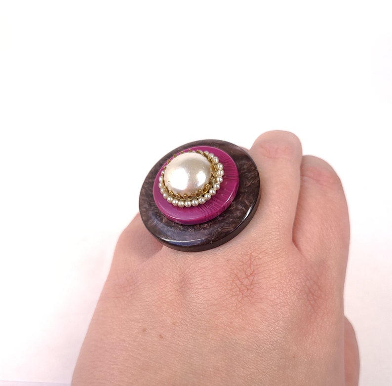 May include: A large, round ring with a brown wooden base and a pink button with a pearl in the center. The ring is worn on a person's finger.