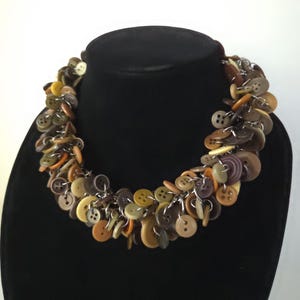 Può includere: Una collana unica composta da bottoni marroni, beige e verde oliva di varie dimensioni. I bottoni sono collegati con catene di metallo color argento, creando un design stratificato e strutturato. La collana è esposta su un manichino nero.