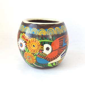 May include: A hand-painted ceramic vase with a brown base, adorned with a vibrant design. The artwork features a red bird, yellow and white flowers, and blue accents. The rim is white, and the vase has a rounded shape.