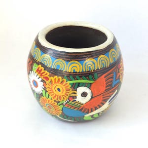 May include: A hand-painted, round ceramic vase with a wide opening. The vase features a colorful floral and bird design with orange, yellow, blue, and white accents. The rim is white, and the interior is dark.