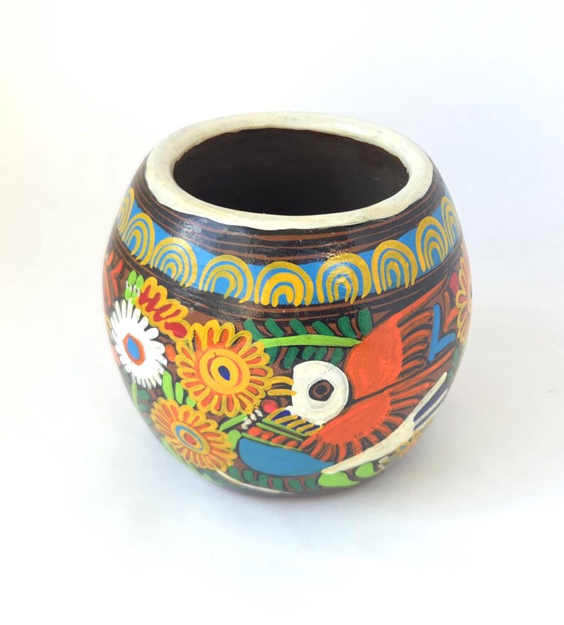 May include: A hand-painted, round ceramic vase with a wide opening. The vase features a colorful floral and bird design with orange, yellow, blue, and white accents. The rim is white, and the interior is dark.