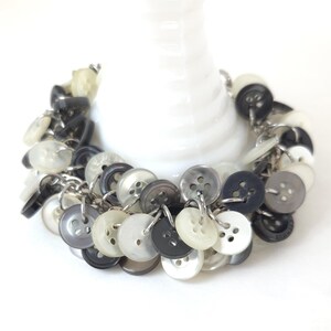 May include: A handmade bracelet featuring a collection of buttons in shades of gray, black, and white. The buttons are attached to a silver chain, creating a unique and eye-catching accessory. The bracelet is displayed on a white background.