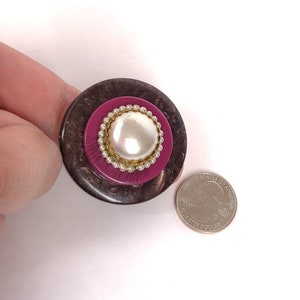 May include: A large, round, brown and pink button with a faux pearl center and a gold beaded trim. The button is about the size of a US quarter.