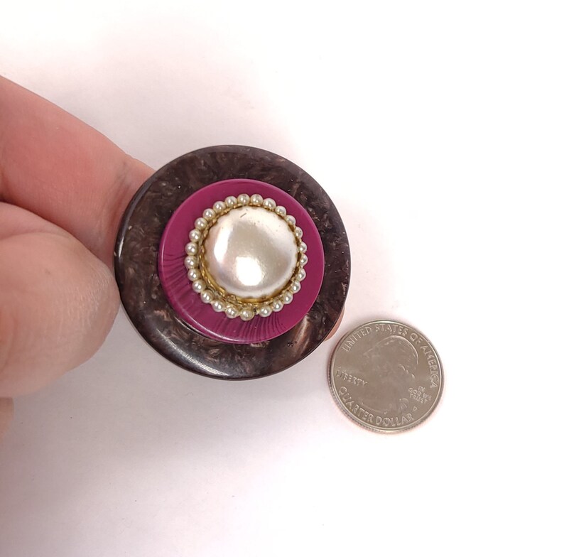 May include: A large, round, brown and pink button with a faux pearl center and a gold beaded trim. The button is about the size of a US quarter.