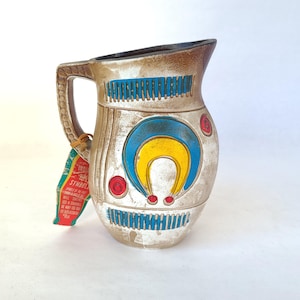 Puede incluir: Una jarra de cerámica con fondo blanco y diseños geométricos coloridos. La jarra tiene un asa y un pico. El diseño presenta una forma de herradura azul, amarilla y roja.