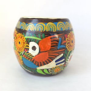 May include: A hand-painted, round ceramic vessel with a brown base, adorned with a vibrant design. The artwork features a colorful bird, flowers, and intricate patterns in orange, blue, yellow, and green.
