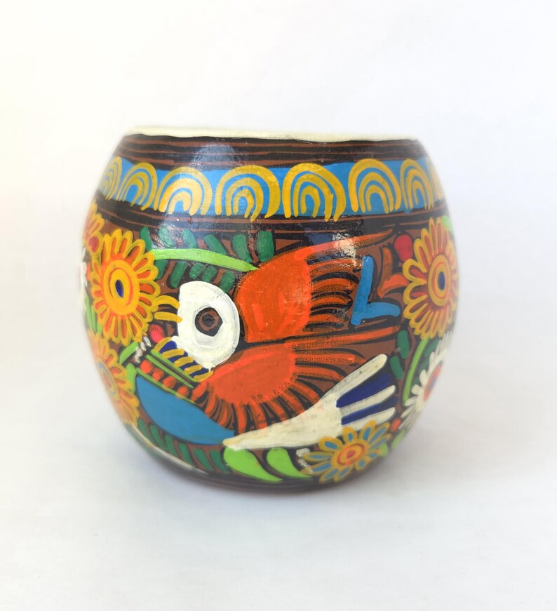 May include: A hand-painted, round ceramic vessel with a brown base, adorned with a vibrant design. The artwork features a colorful bird, flowers, and intricate patterns in orange, blue, yellow, and green.
