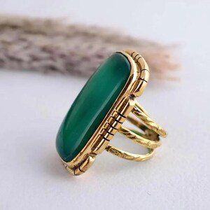 May include: A gold-coloured ring with a large, oval green stone. The ring has a detailed, textured band and a decorative frame around the stone. The design is vintage-inspired and eye-catching.