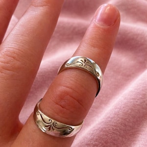 Könnte beinhalten: Zwei silberne Ringe mit einem dekorativen floralen Design werden auf einem Finger gezeigt. Die Ringe haben eine polierte Oberfläche und eine gebogene Form. Der Hintergrund ist ein weicher rosa Stoff.