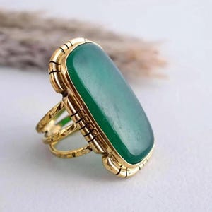 May include: A gold-coloured ring with a large, rectangular, emerald-green stone. The ring has an ornate, textured band and a decorative bezel around the stone. The design is vintage-inspired, with a bold and eye-catching aesthetic.