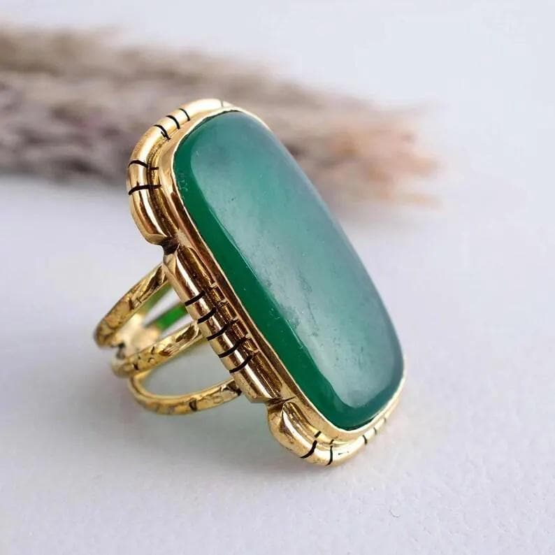 May include: A gold-coloured ring with a large, rectangular, emerald-green stone. The ring has an ornate, textured band and a decorative bezel around the stone. The design is vintage-inspired, with a bold and eye-catching aesthetic.