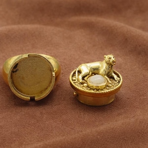 Peut inclure: Deux composants métalliques dorés. L'un est une bague avec un sertissage plat et circulaire. L'autre est un petit récipient rond avec une figurine de tigre en métal doré détaillée sur le dessus et une pierre blanche au centre.
