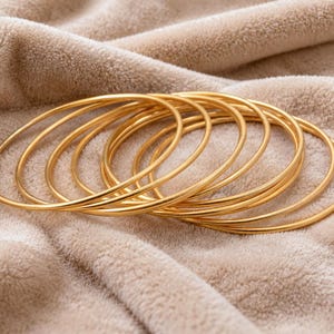 May include: A set of seven thin, circular gold-coloured bangles arranged in a slightly overlapping stack. Each bangle features a smooth, polished, and continuous metal surface with a consistent round profile and a bright, reflective yellow-gold finish.