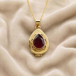 May include: A gold-colored pendant necklace with a teardrop-shaped locket. The locket features a dark red gemstone surrounded by a decorative gold border. The necklace chain is also gold.
