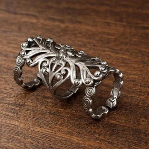 Puede incluir: Un anillo de dedo articulado de color plateado con un diseño floral ornamentado. El anillo presenta tres bandas conectadas, cada una con intrincados volutas y elementos decorativos. El anillo es de metal y está sobre una superficie de madera.