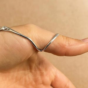 May include: A silver-colored ring with a V-shaped design. The ring is worn on a finger, with the V shape extending across the top of the finger. The ring appears to be made of a shiny metal, reflecting light.