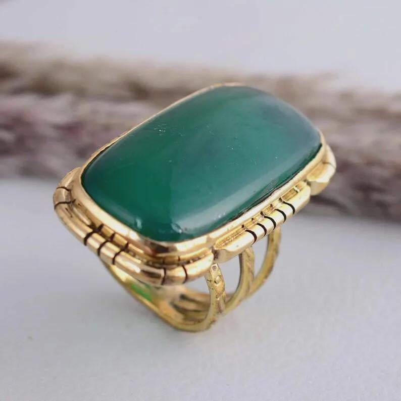 May include: A gold ring with a large, rectangular, emerald-green stone. The ring has a detailed, textured band and a decorative setting around the stone. The ring is a statement piece, suitable for special occasions.
