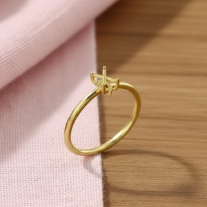May include: A gold-colored ring with a prong setting. The ring is set against a pink fabric and a wooden surface. The ring's band is thin and circular, and the setting is designed to hold a gemstone.