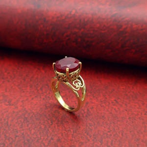 Op de afbeelding: Een gouden ring met een grote, ovaal geslepen, dieprode edelsteen. De ring heeft een sierlijk filigraanontwerp op de band en rond de edelsteen setting. De ring staat tegen een rode achtergrond.