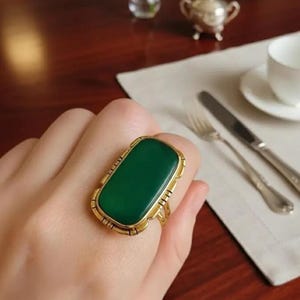 May include: A gold ring with a large, rectangular, emerald-green stone. The ring has an ornate, detailed setting. The ring is on a person's finger, with a blurred background including a table setting.