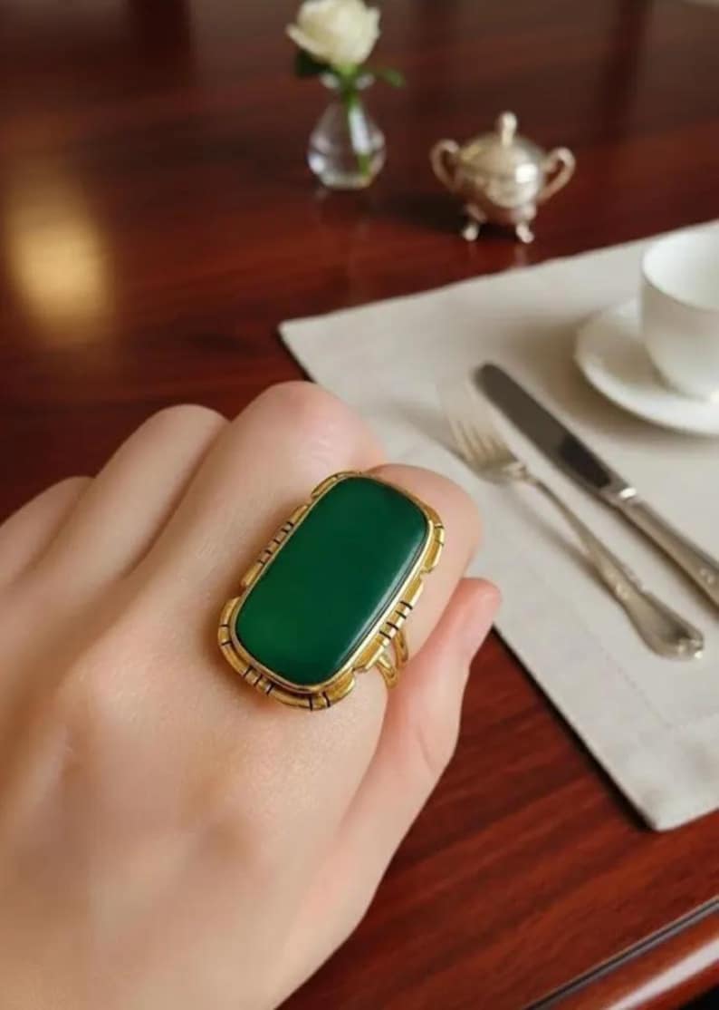 May include: A gold ring with a large, rectangular, emerald-green stone. The ring has an ornate, detailed setting. The ring is on a person's finger, with a blurred background including a table setting.