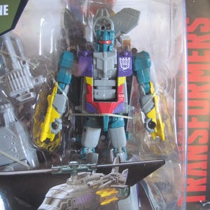 May include: A Transformers action figure of Vortex, a blue, gray, yellow, and purple robot that transforms into a helicopter. The figure is packaged in a clear plastic blister pack with a black and white background. The packaging includes the Transformers logo and the text "Ages 8+".